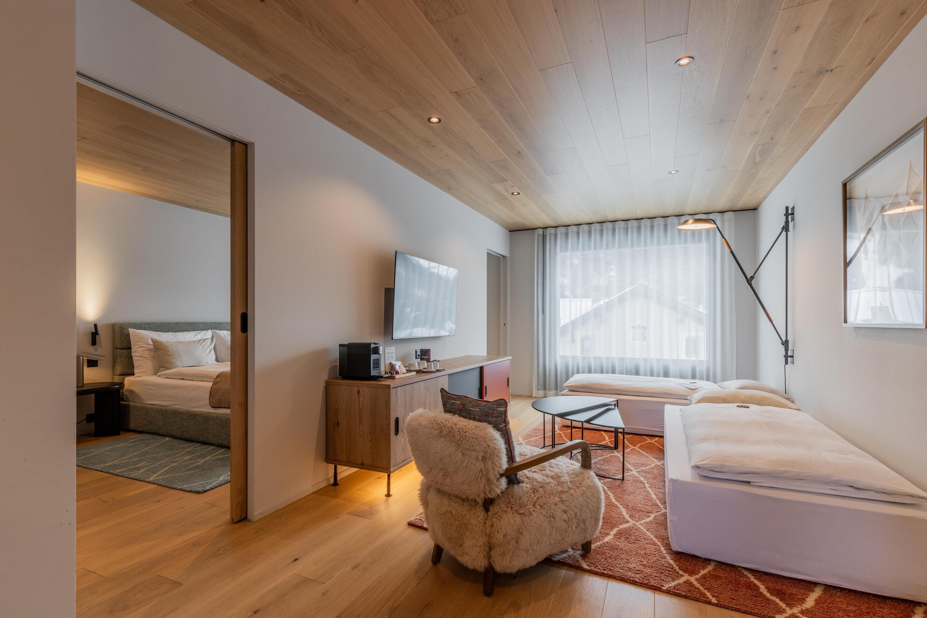A modern hotel room with a wooden ceiling, a bed, a sofa, a desk with a TV, and a large window with curtains.