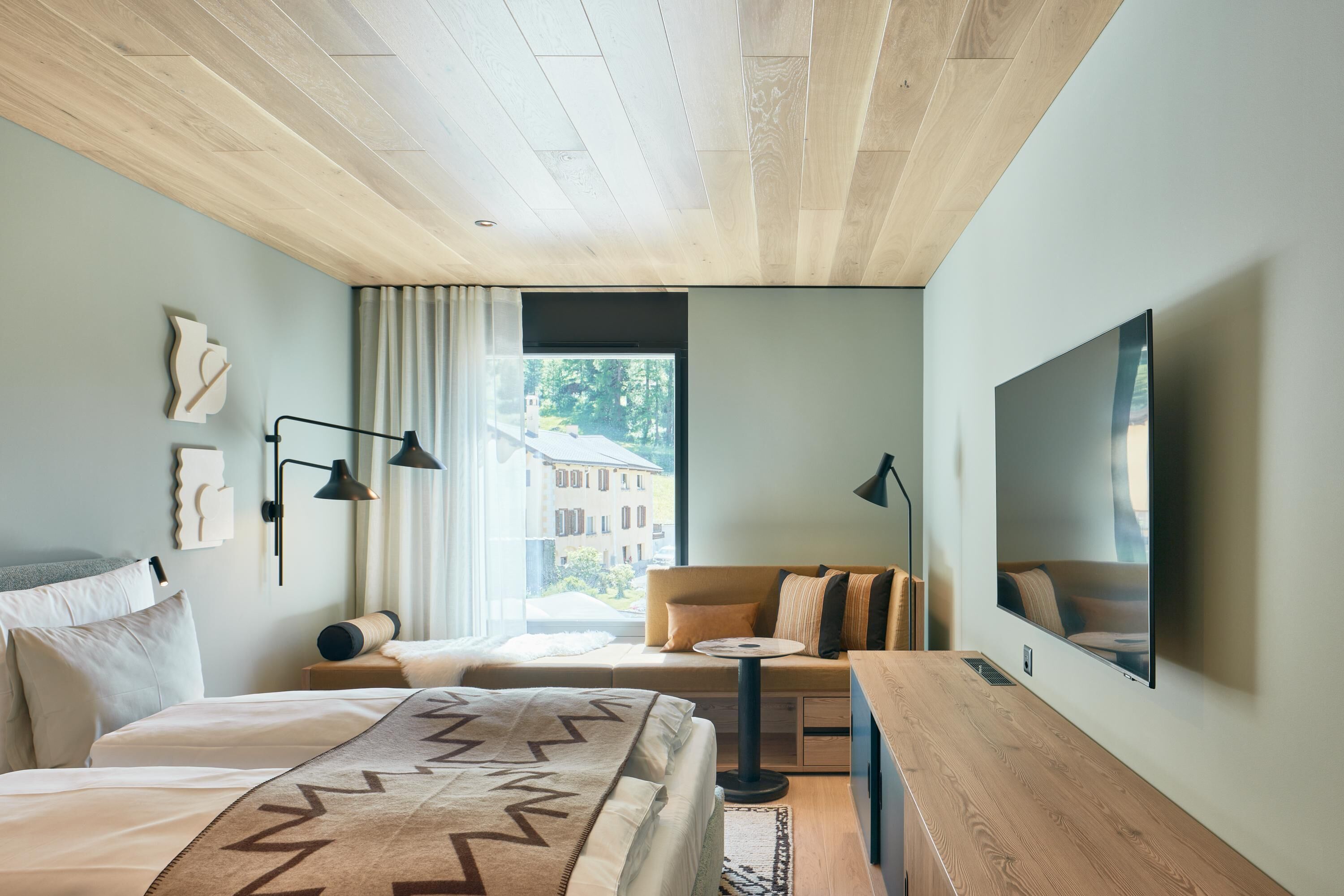A modern hotel room with a large bed, a sofa, a small table, and a TV mounted on the wall.