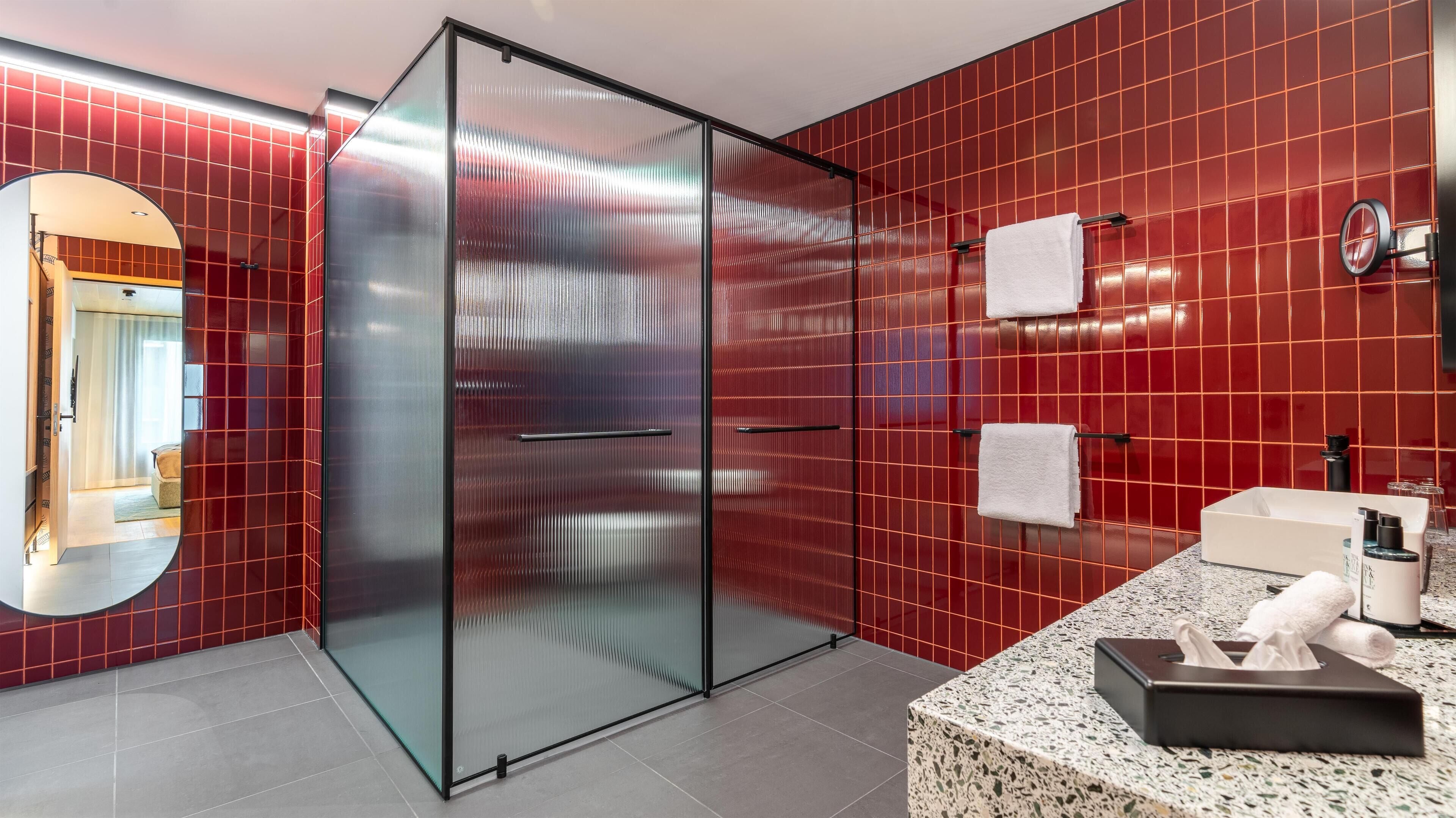 A modern bathroom with red tiles, a glass shower enclosure, a granite countertop, and a mirror.