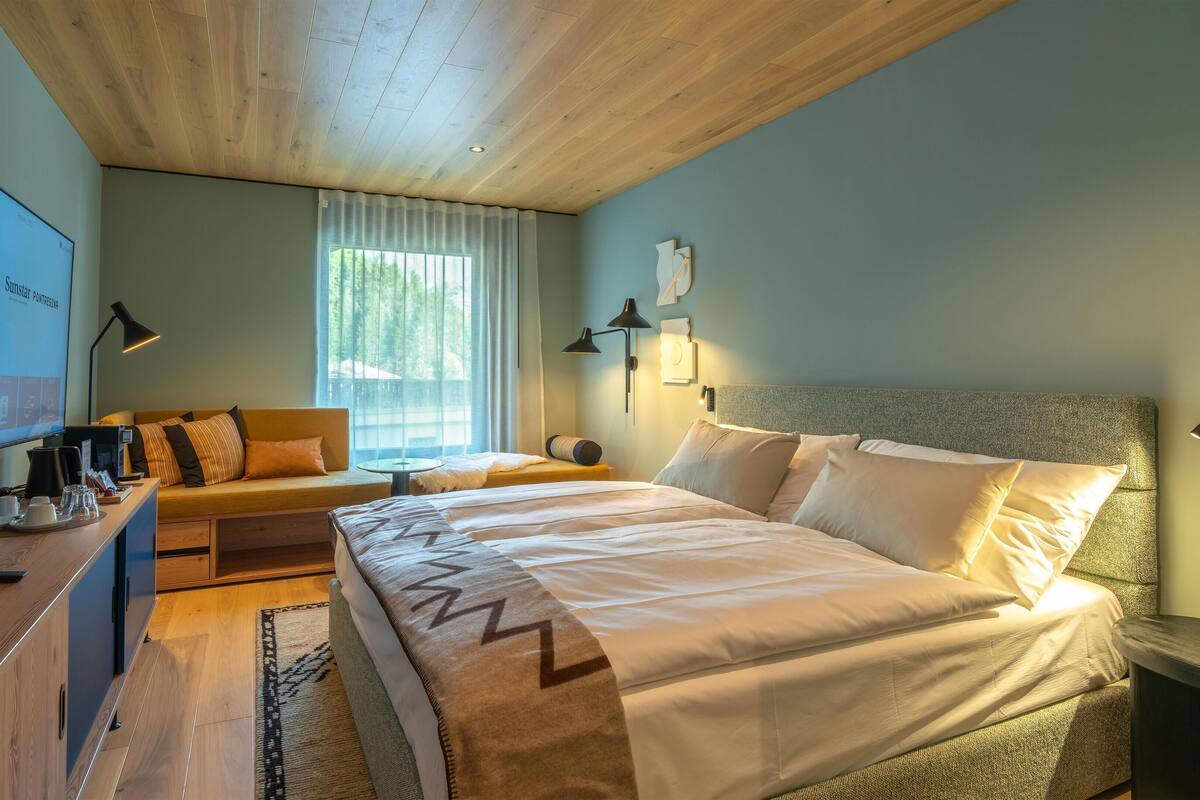 Ein modernes Hotelzimmer mit Bett, einer Sitzecke, einem Schreibtisch mit Kaffeemaschine und einem Fernseher, der eine Präsentation zeigt.