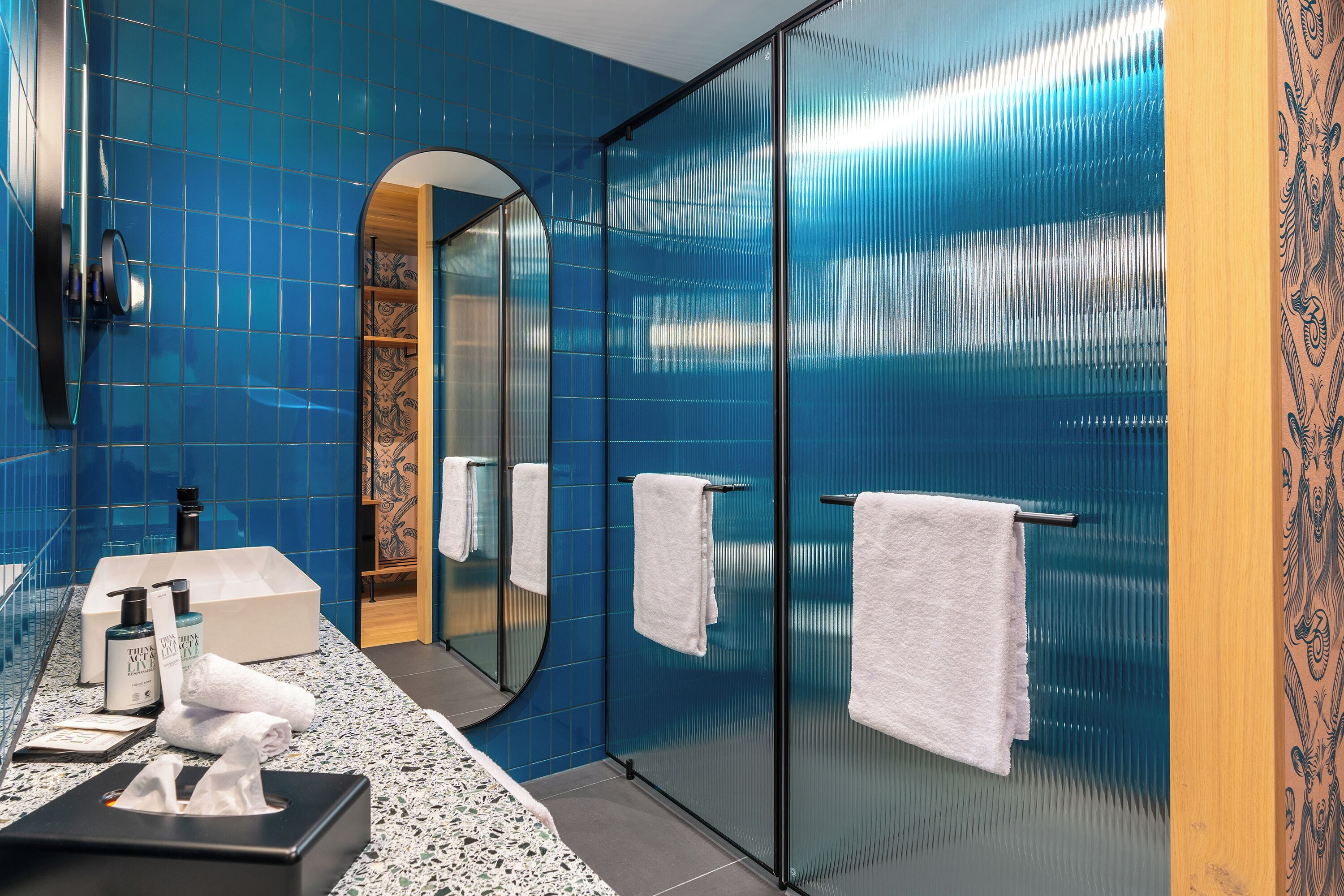 A modern bathroom with blue tiles, a large mirror, a sink with toiletries, and a glass shower enclosure.