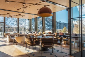 A modern lounge with leather sofas and chairs, large windows offering mountain views, and a wooden ceiling with pendant lighting. - Sunstar Pontresina (Pontresina)