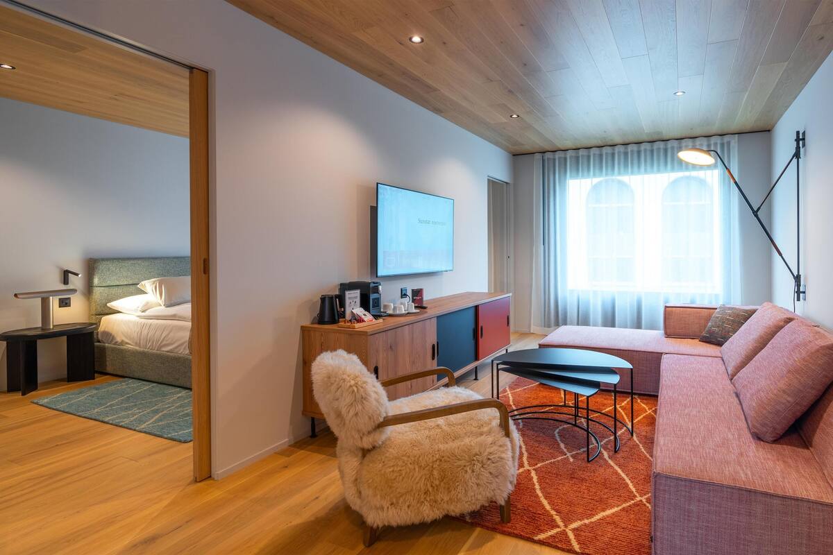Ein modernes Wohnzimmer mit einer Couch, einem kleinen Tisch, einem gemütlichen Sessel, einem Fernseher und einer Holzbalkendecke.