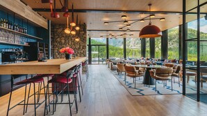 Uma área de bar moderna com balcão de madeira, banquetas vermelhas e parede de pedra. Há várias luminárias pendentes e uma grande janela com vista para o exterior.