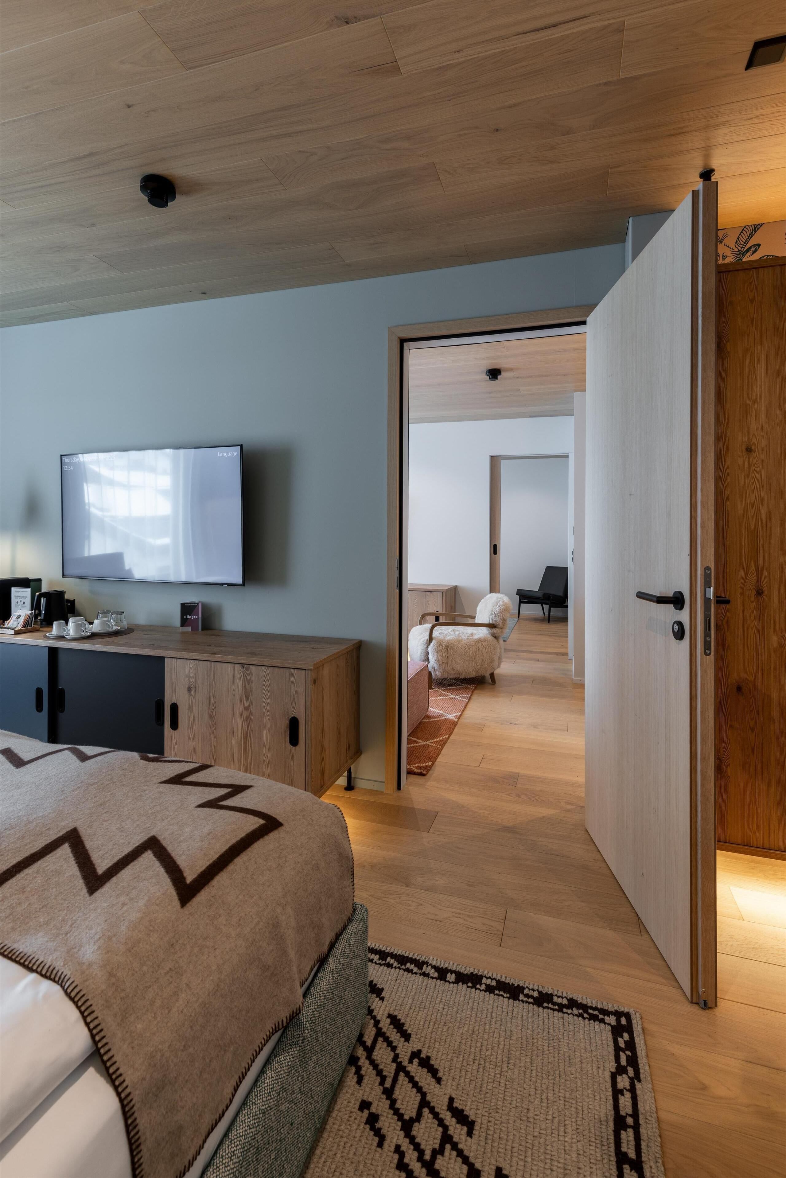 Un dormitorio moderno con techo de madera, una cama grande con colcha estampada, un televisor sobre un mueble de madera y vista a una habitación contigua con una silla y un sofá.