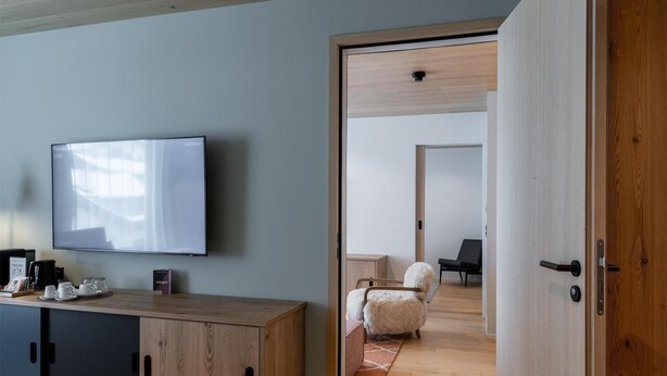 Un dormitorio moderno con techo de madera, una cama grande con colcha estampada, un televisor sobre un mueble de madera y vista a una habitación contigua con una silla y un sofá.
