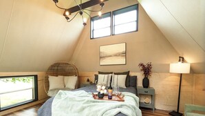 A cozy bedroom with a large bed, a bedside table, a lamp, and a view of the outdoors. - Goat Farm Tree House on Creek (Newport)