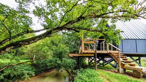 A wooden deck with a staircase leading to a house built on stilts over a river. - Goat Farm Tree House on Creek (Newport)