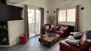 Ein Wohnzimmer mit einem roten Sofa, einem roten Sessel, einem Glastisch und einem roten Hocker. An der Wand ist ein Flachbildfernseher angebracht, es gibt eine Lampe und ein Fenster mit Vorhängen.