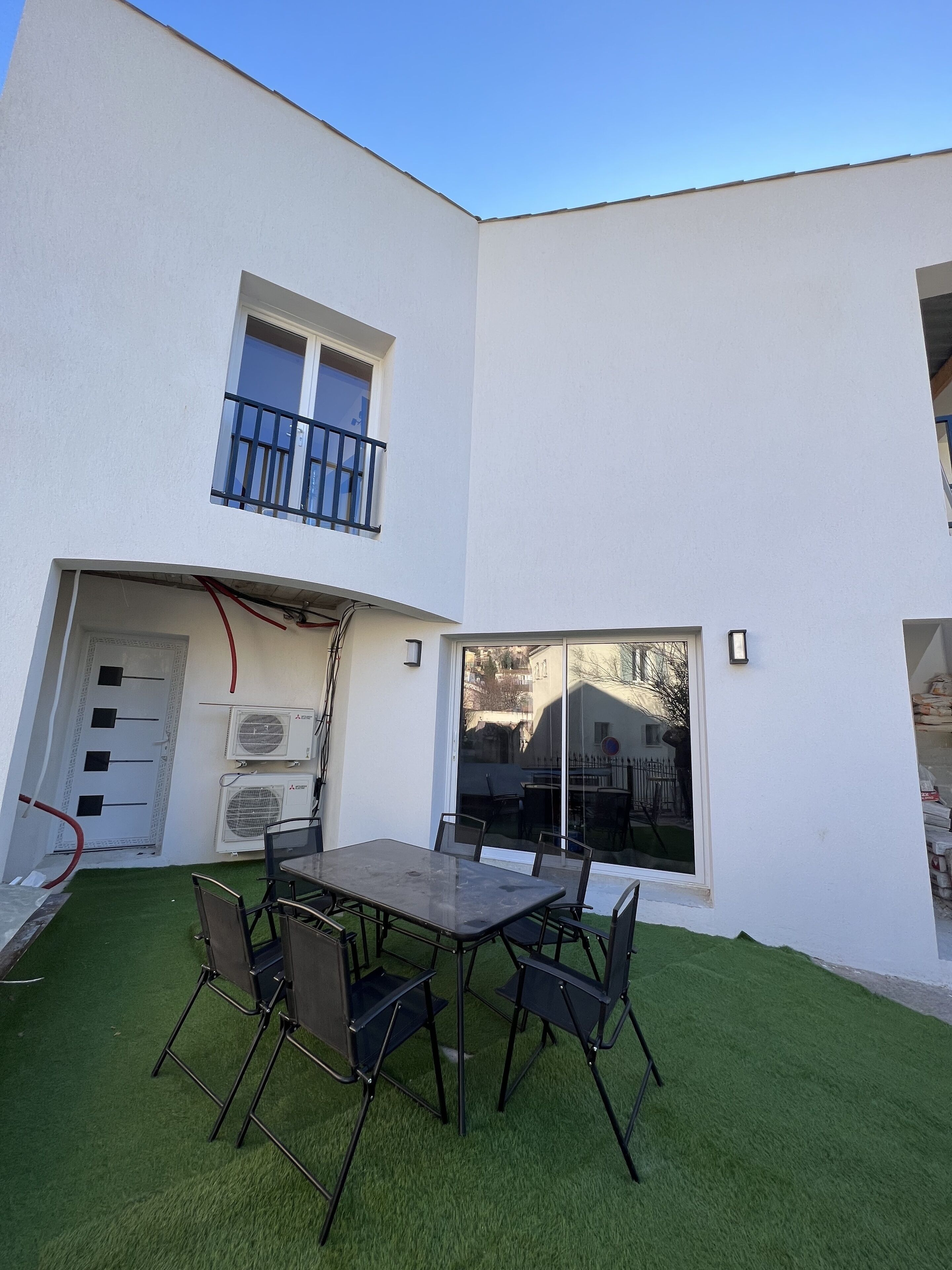 Un edificio moderno con techo plano, un balcón con barandillas negras y un vidrio que da a un patio con mesa y sillas.