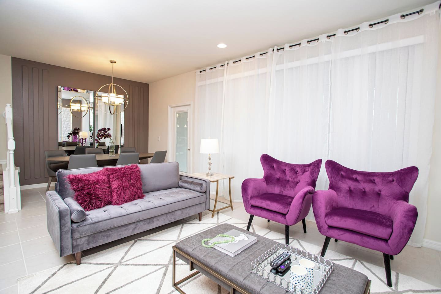 A modern living room with a grey sofa, two purple armchairs, a coffee table, and a dining area in the background.