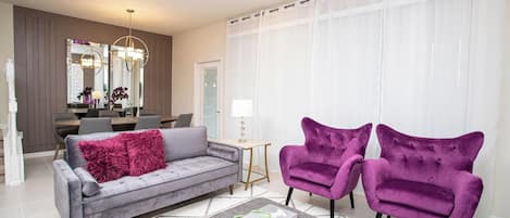A modern living room with a grey sofa, two purple armchairs, a coffee table, and a dining area in the background.