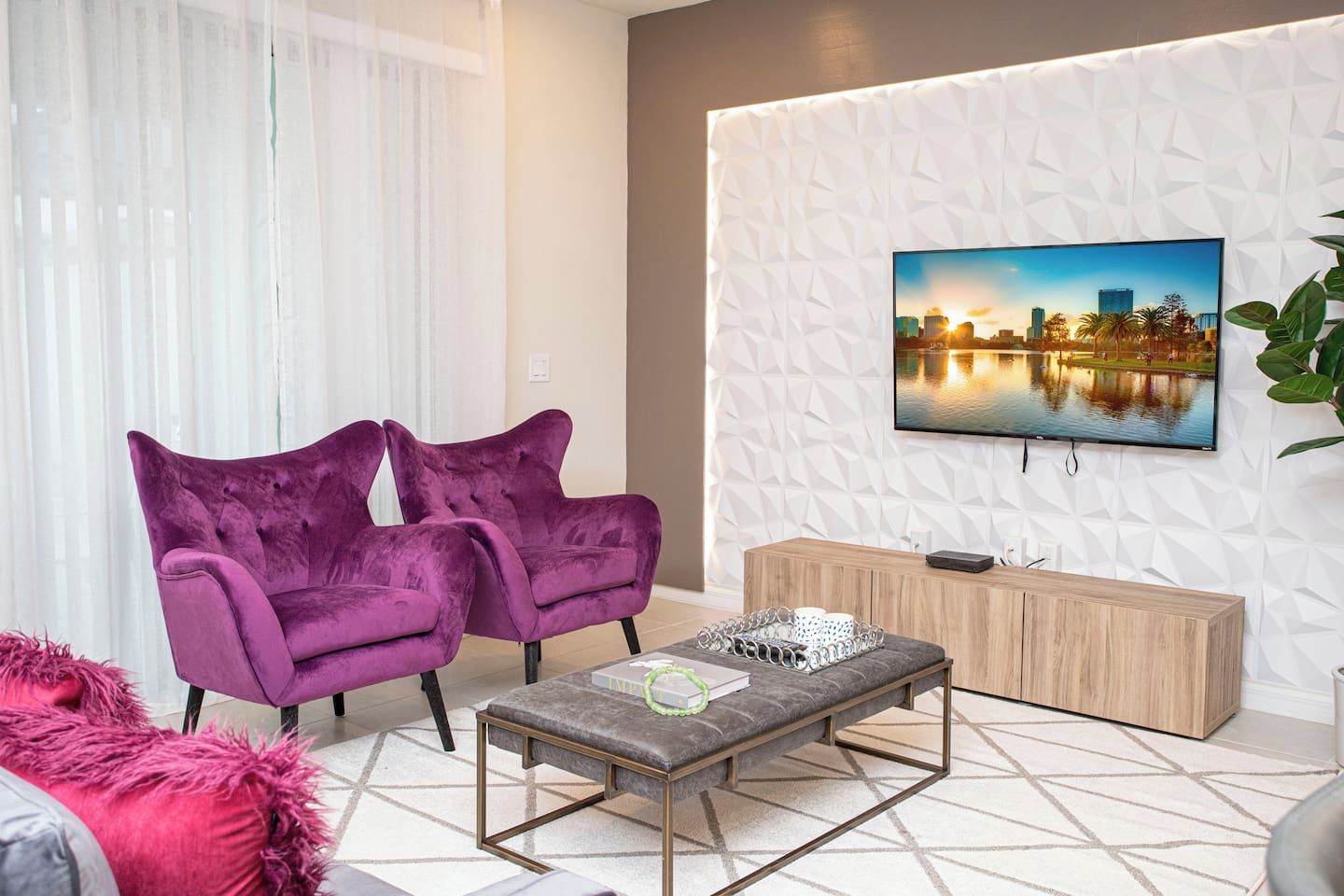 A modern living room with two purple armchairs, a grey ottoman, a wooden sideboard, and a flat-screen TV mounted on a textured wall.