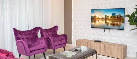 Un salon moderne avec deux fauteuils violets, un pouf gris, un buffet en bois et un téléviseur à écran plat fixé sur un mur à texture.