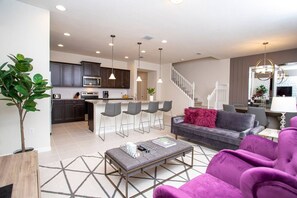 Ein modernes Wohnzimmer mit einem grauen Sofa, einem Couchtisch und einem violetten Sessel. Es gibt eine Küchenzeile mit dunklen Schränken und eine Essecke mit Barhocker.