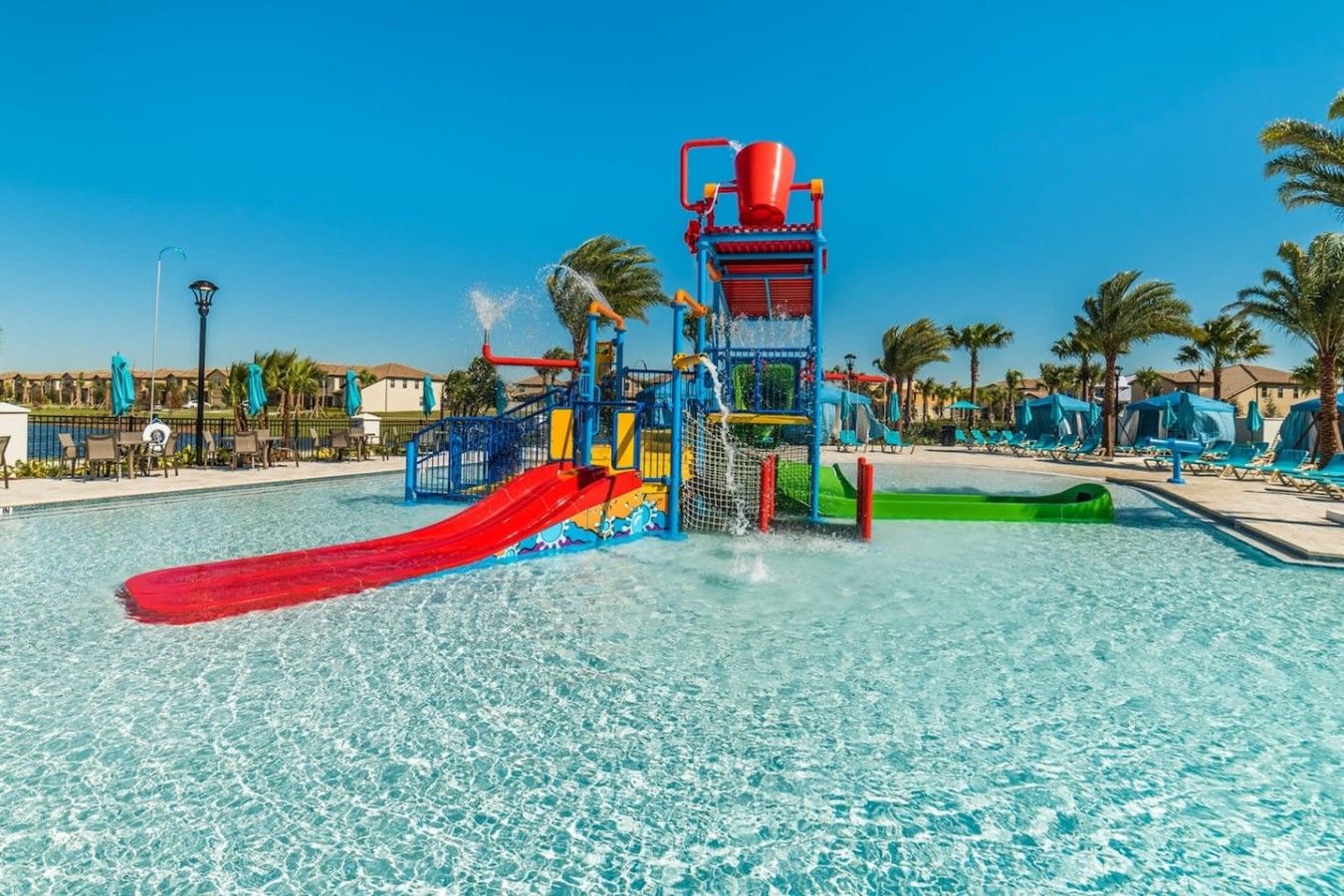 A water park with a red slide, a water play structure, and a pool area with lounge chairs and umbrellas.