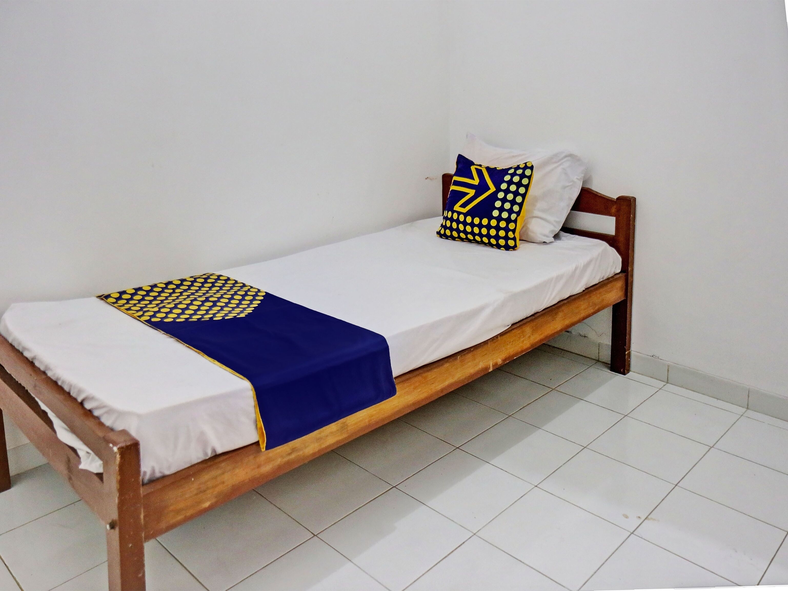 Een eenpersoonsbed met houten frame, wit beddengoed en een deken met blauw-geel patroon.