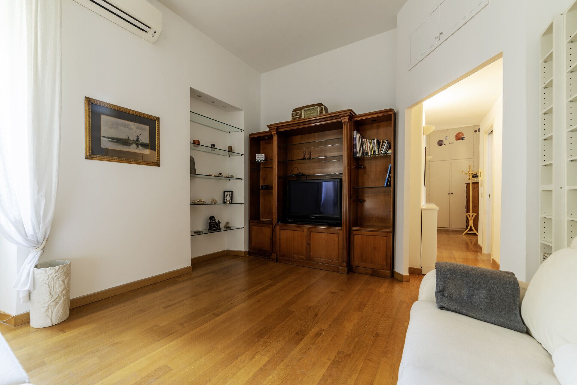 A living room with a wooden TV stand, a white sofa, and a glass shelving unit.