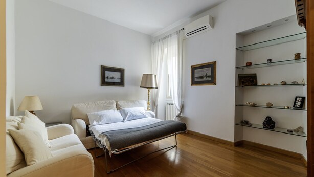 Une chambre d’hôtel comprenant un lit, une chaise, une lampe, une fenêtre avec des rideaux et une étagère ornée d’objets décoratifs.