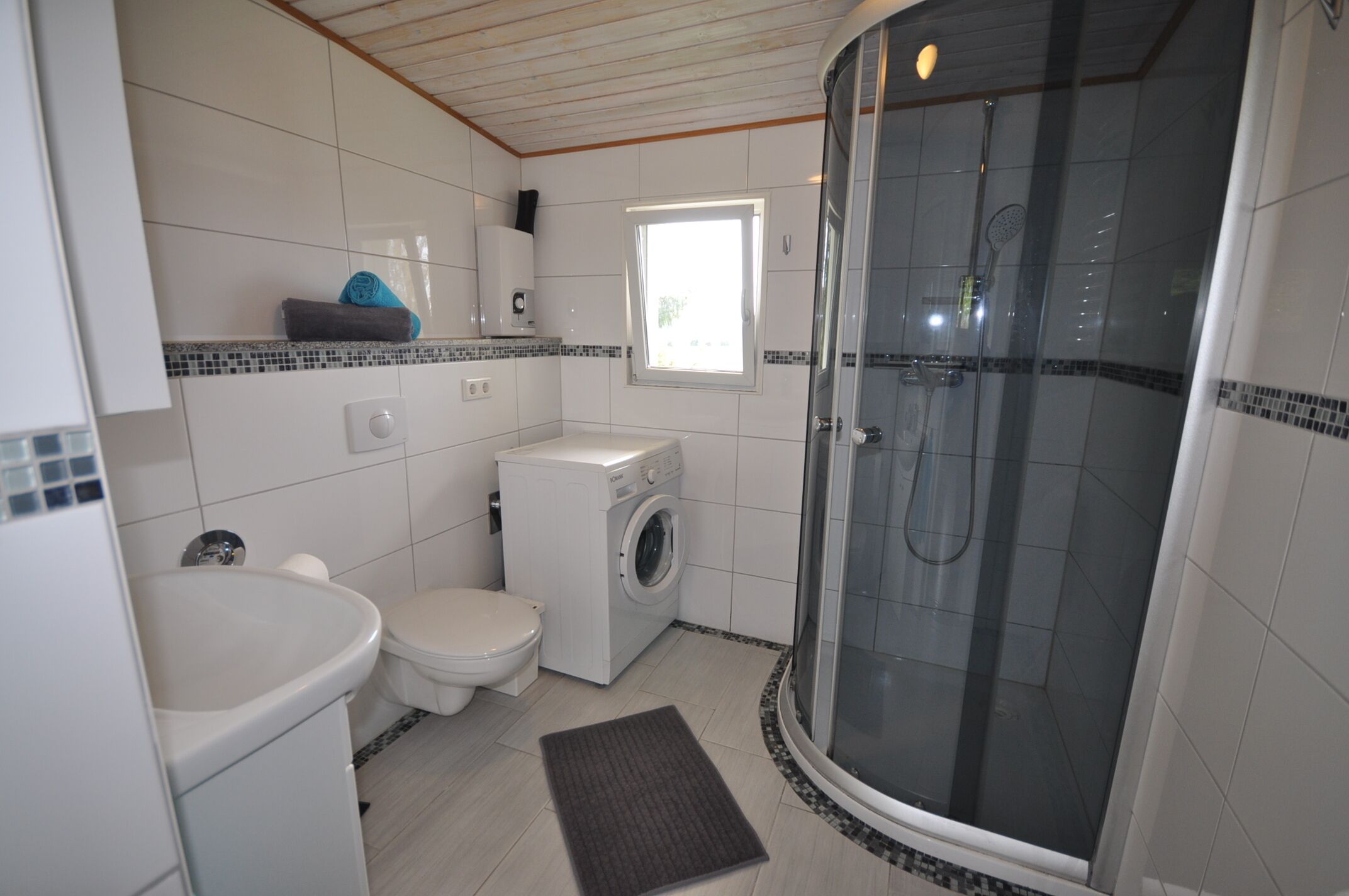 Une petite salle de bain comprenant des toilettes, un lave-linge et une douche fermée par des parois en verre.