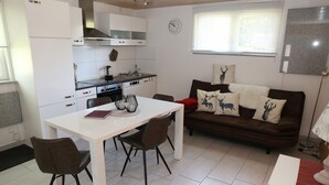 A compact living space with a kitchenette, a dining table with chairs, a brown sofa with decorative pillows, and a clock on the wall. - Vacation home Beerberg, cozy vacation home at the biosphere reserve Thuringian Forest (Suhl)