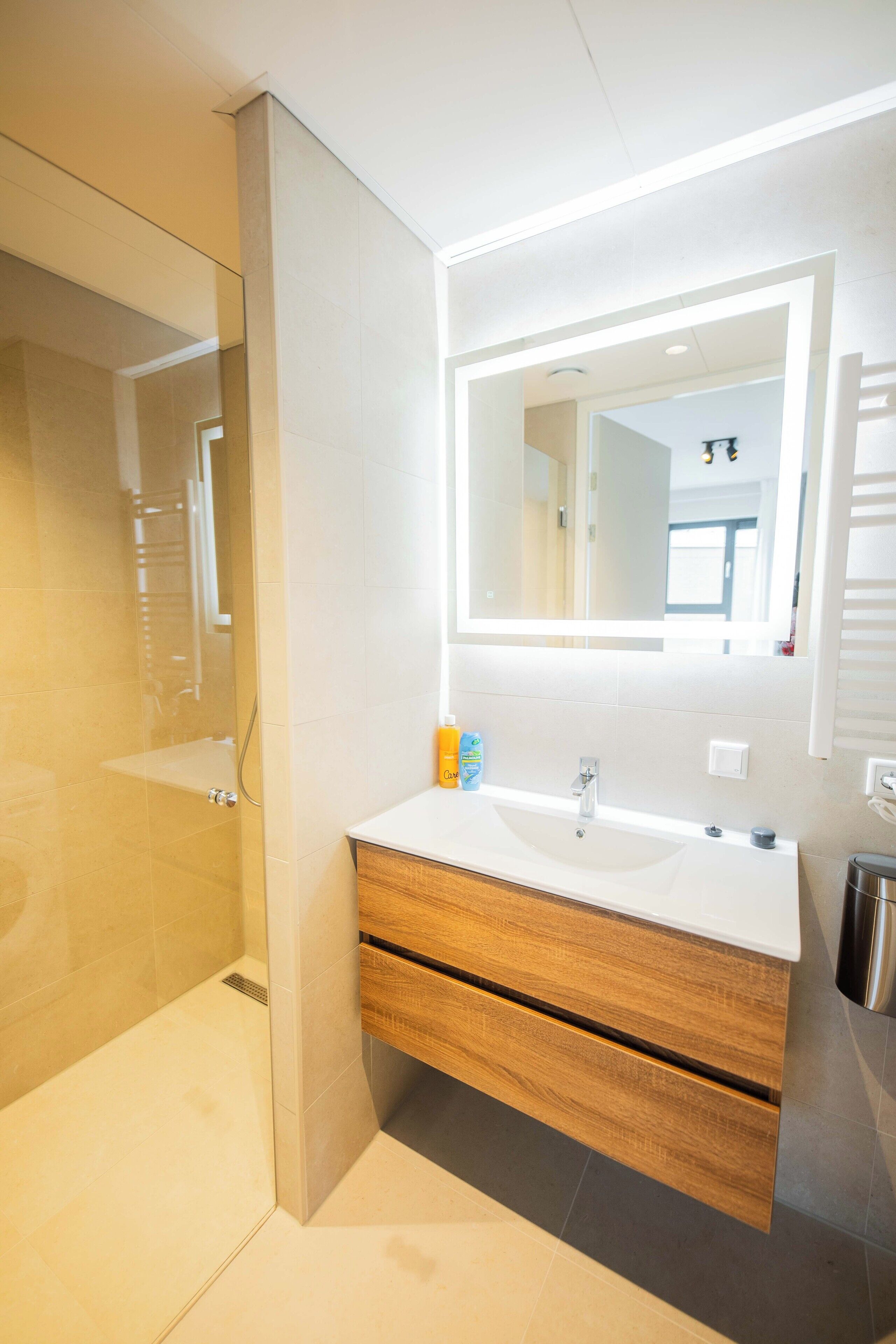Une salle de bain moderne dotée d’une douche à parois de verre, d’un lavabo blanc avec un meuble-vasque en bois et d’un grand miroir avec éclairage intégré.