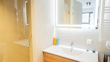 Une salle de bain moderne dotée d’une douche à parois de verre, d’un lavabo blanc avec un meuble-vasque en bois et d’un grand miroir avec éclairage intégré.