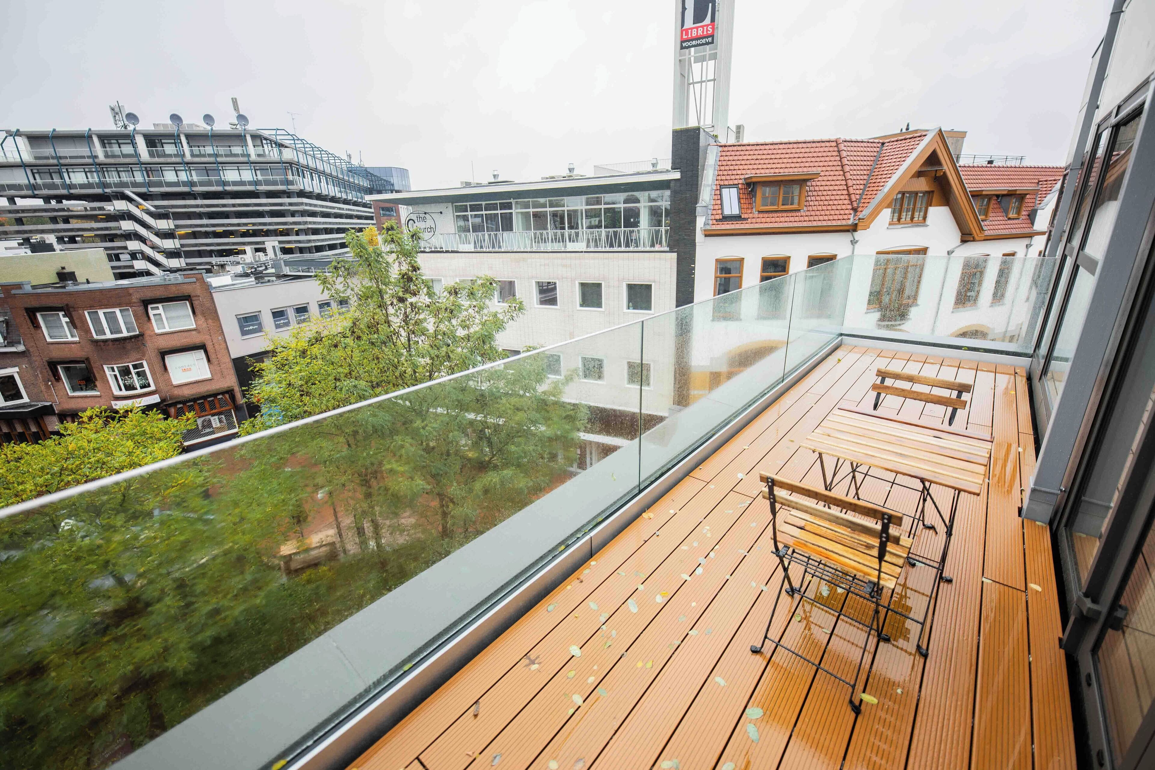Een balkon met houten vloer en metalen hekwerk, met uitzicht op een stadsgezicht met diverse gebouwen en een boom.