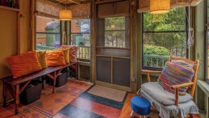 A cozy interior with a wooden bench, a chair with a colorful cushion, and a view of greenery outside through large windows.