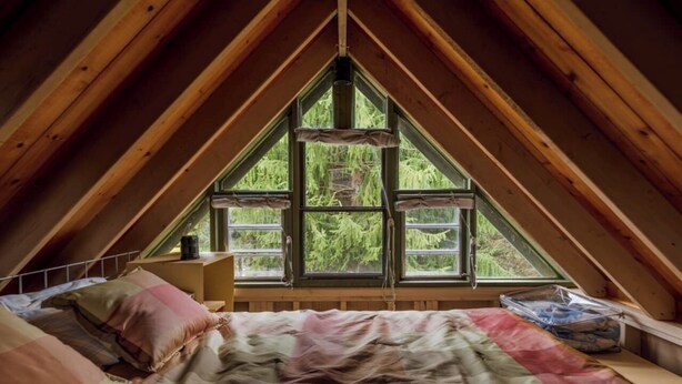 A bedroom with a sloped ceiling, a bed with a patterned bedspread, a nightstand, and a window with a view of greenery.