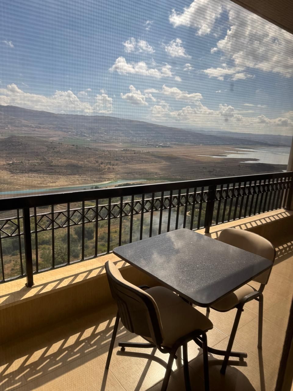 A balcony with a table and chairs, overlooking a landscape with a river and distant hills.