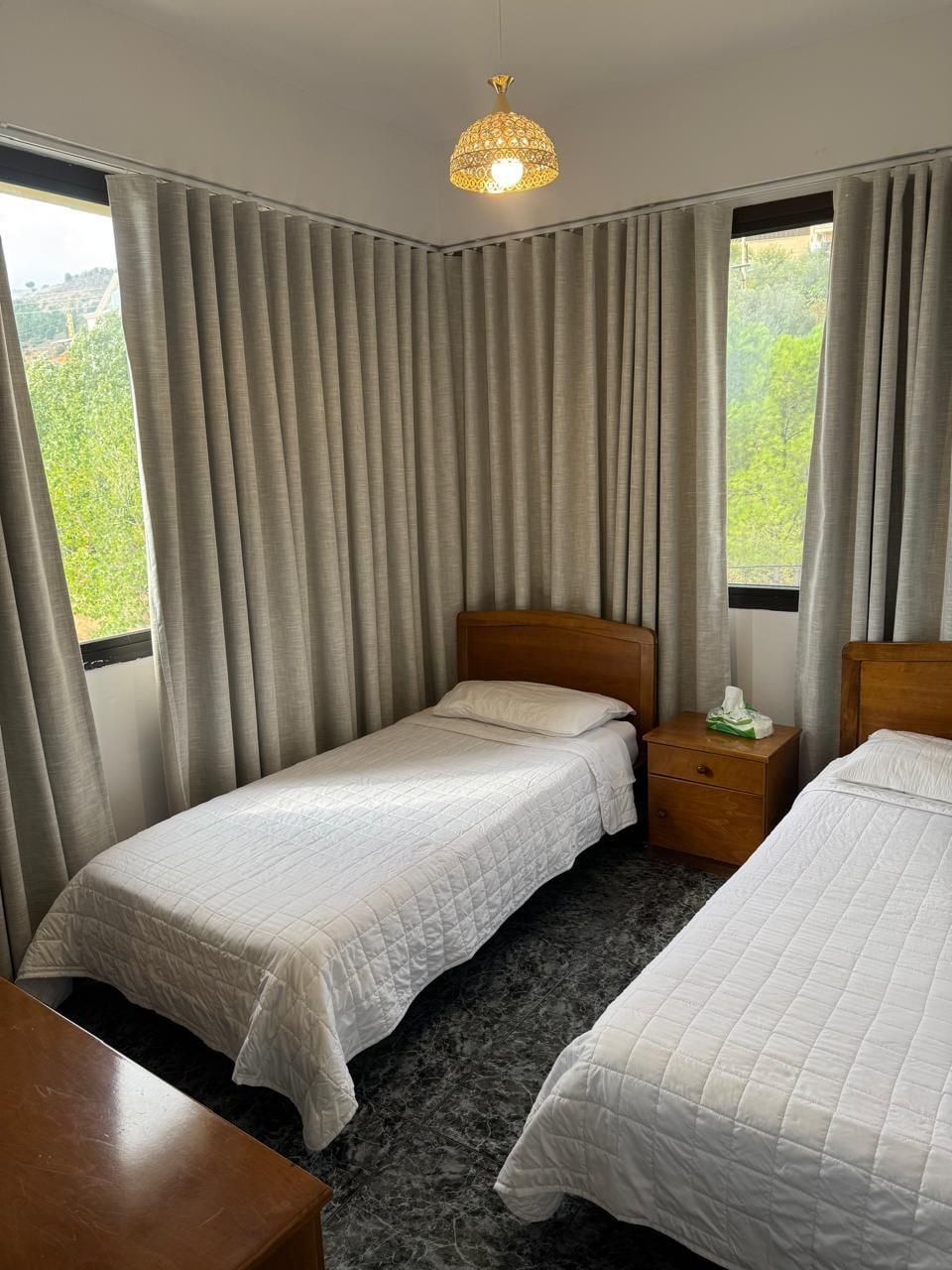 Habitación de hotel con dos camas, una mesita de noche de madera, una lámpara y vistas a la vegetación a través de las ventanas.