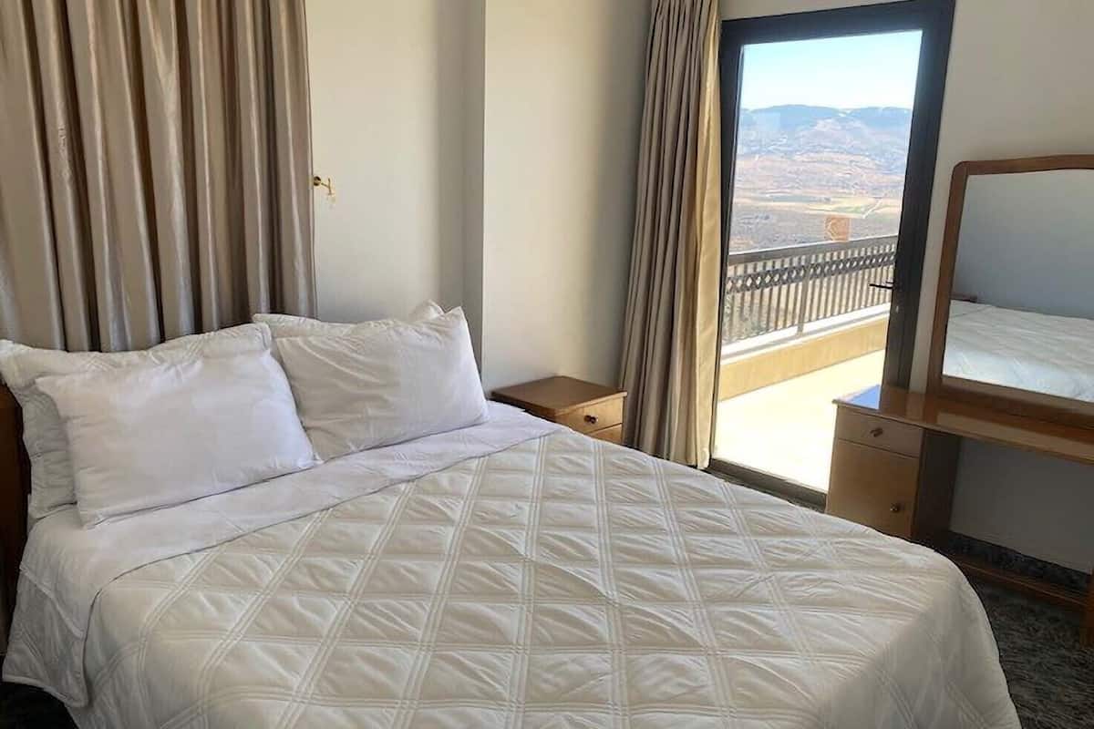 A hotel room with a bed, a nightstand, a dresser, and a balcony with a view of the landscape.