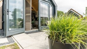 A modern house with a glass door entrance, a concrete patio, and a large potted plant in the foreground. - Sun, sea, sand, wind, marram grass – this 4-person chalet embodies the pure beach feeling from your (Burgh-Haamstede)