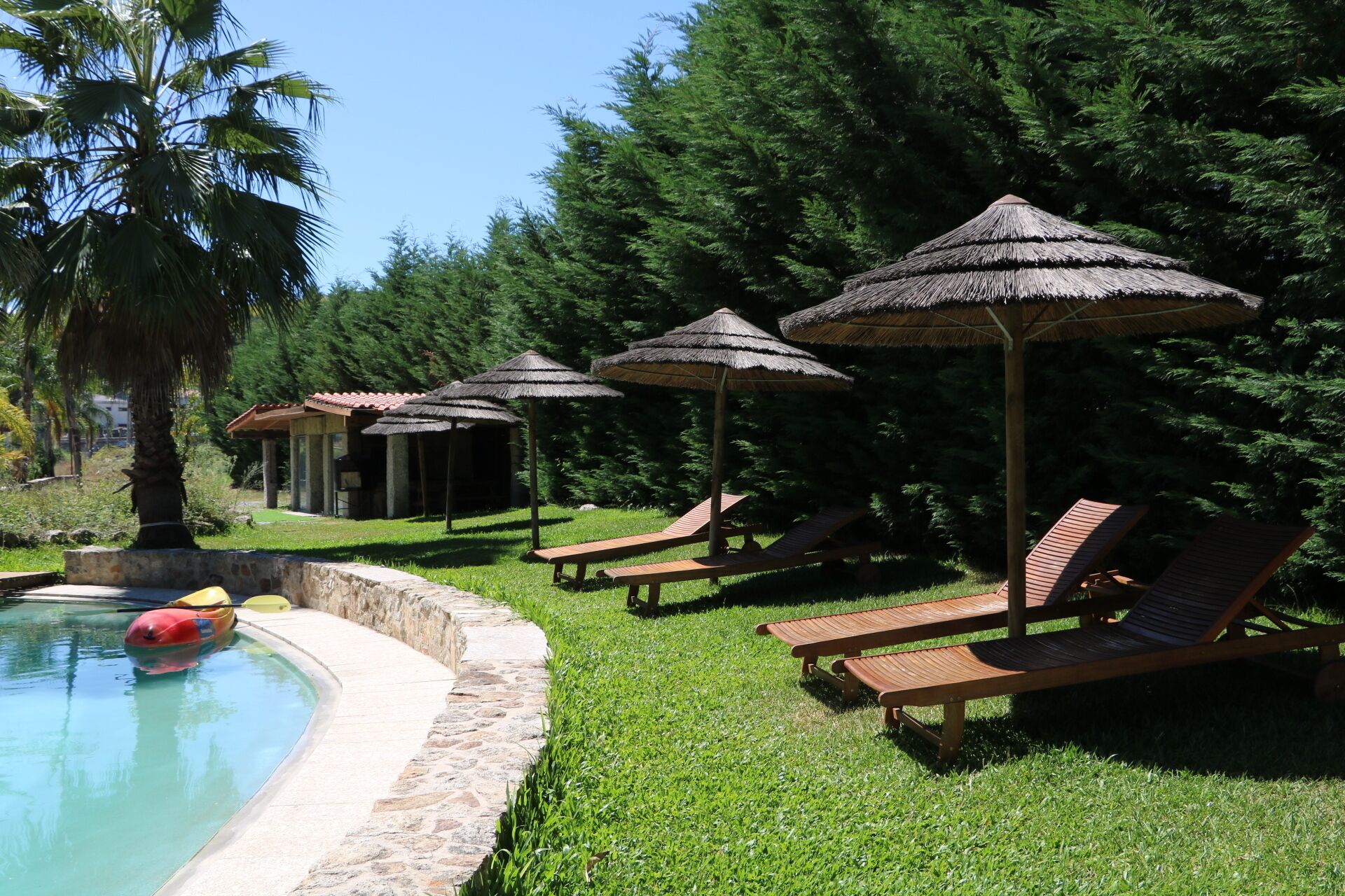 A pool area with lounge chairs and thatched umbrellas, surrounded by greenery and a small building.