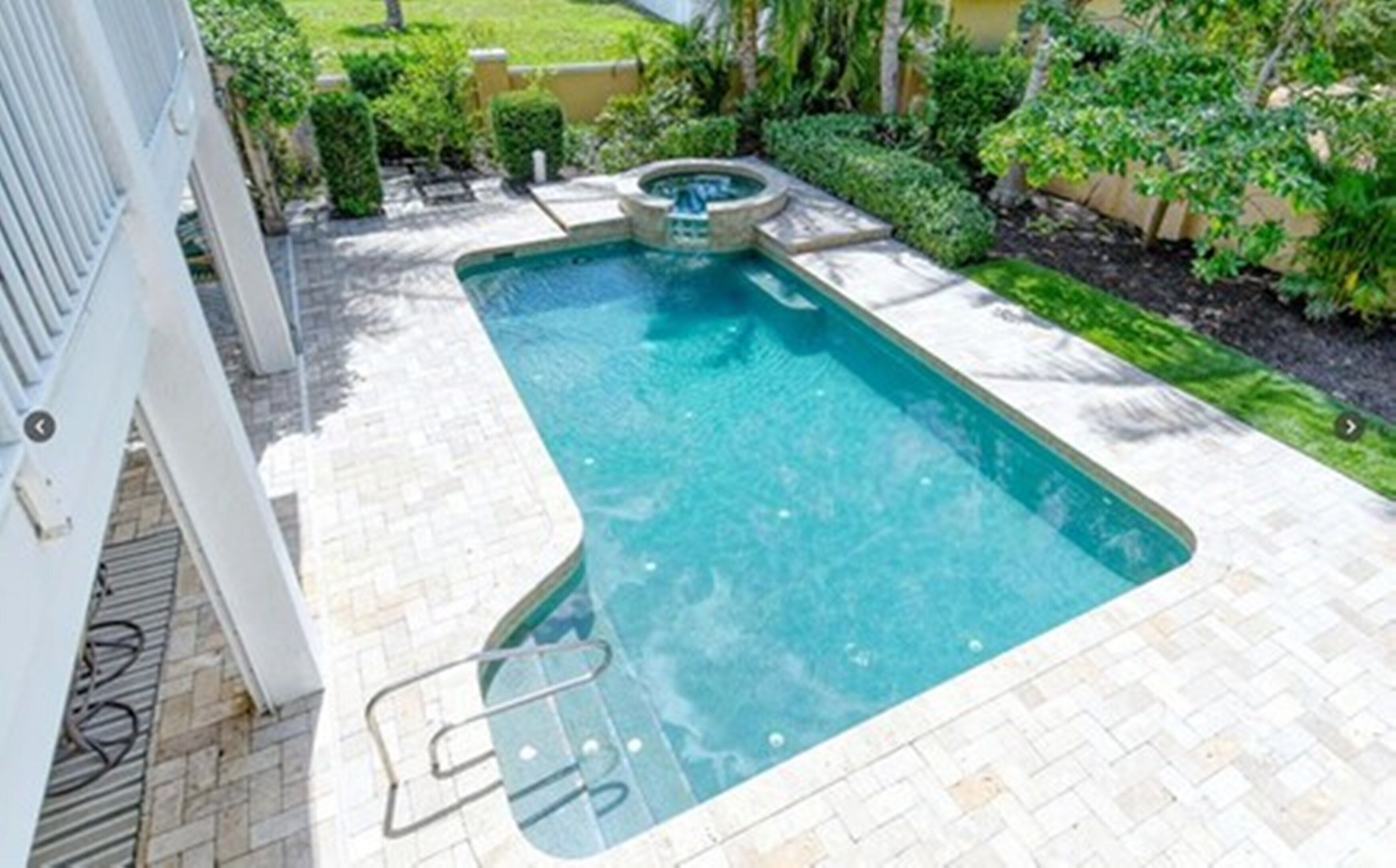 A rectangular swimming pool with a hot tub, surrounded by a paved deck and greenery.