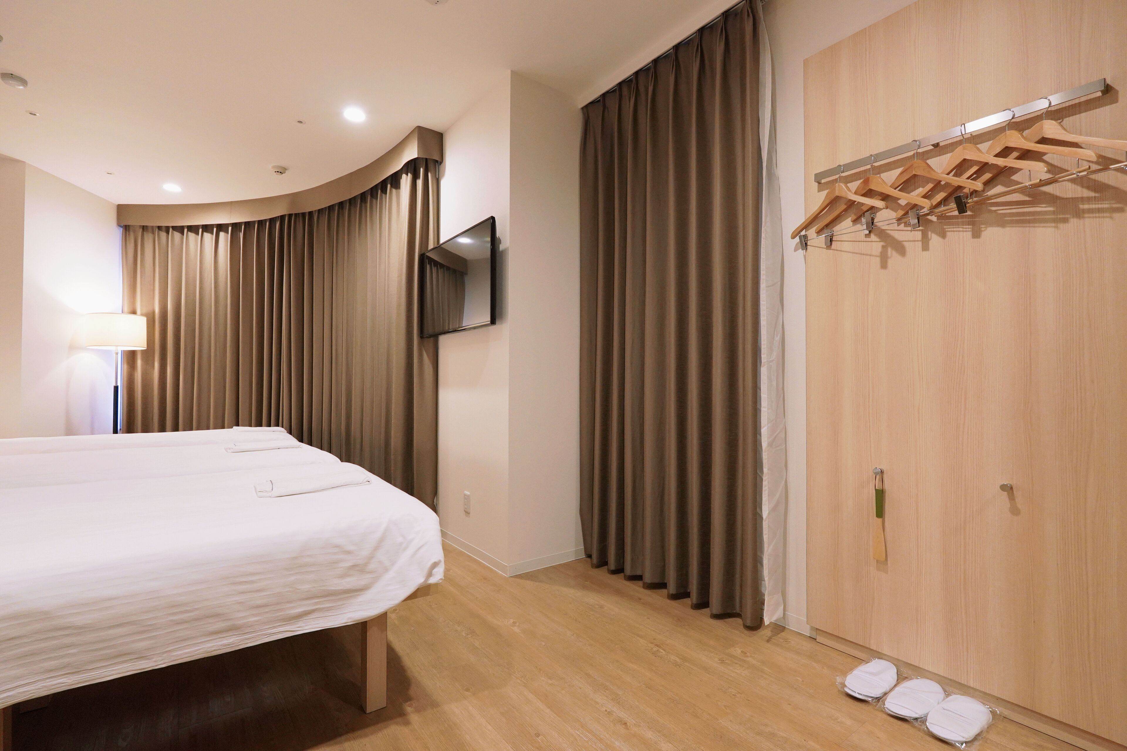 A hotel room with a bed, a TV mounted on the wall, a wooden wardrobe, and a clothes drying rack.