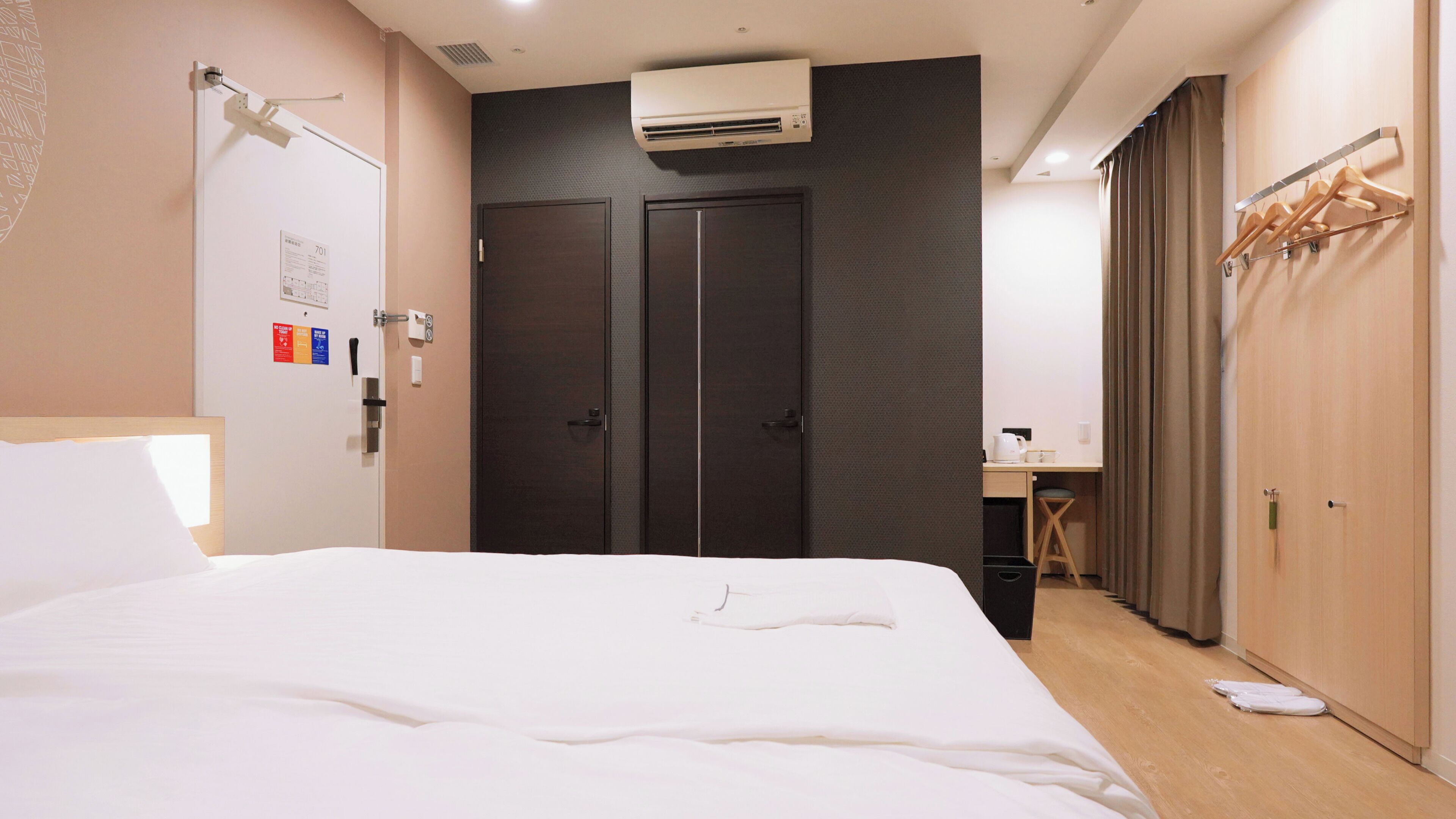 A hotel room with a bed, a wardrobe, a desk with a chair, and an air conditioning unit.