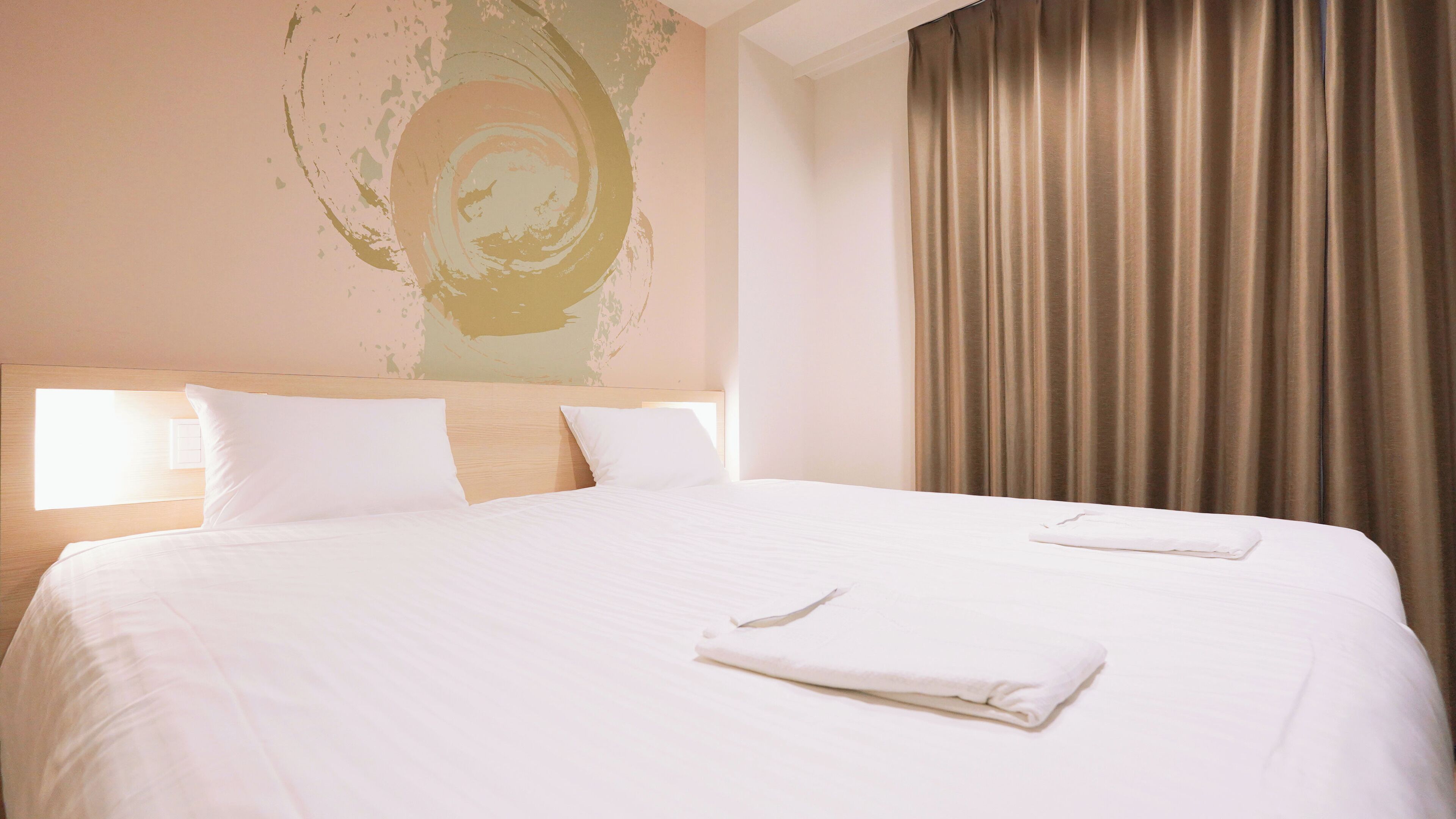 A hotel room with a large bed, two pillows, a headboard, a wall with a circular design, and a curtain.