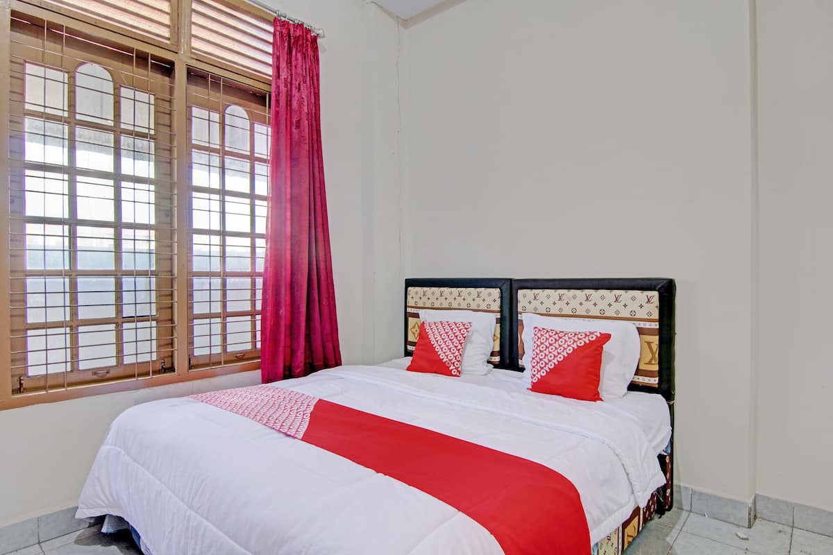 A neatly made bed with a white and red bedspread, a headboard with a patterned design, and a window with wooden blinds and a red curtain.