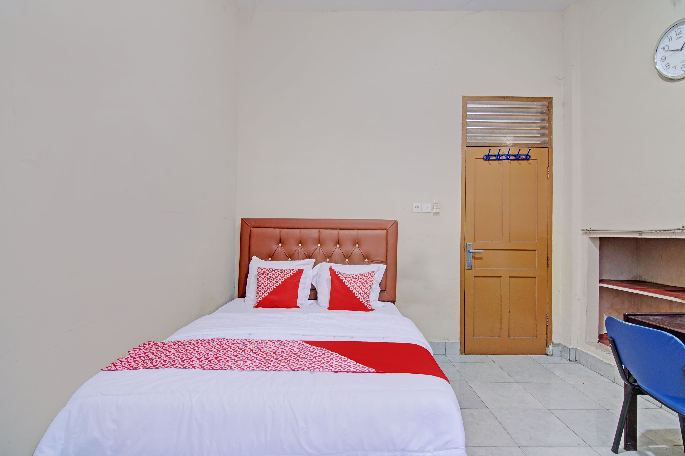 A bedroom with a bed, a wooden door, a blue chair, and a clock on the wall.