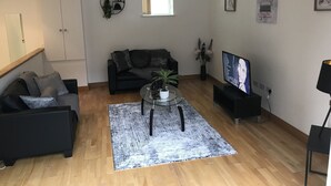 Un soggiorno moderno con un divano nero, un tavolino in vetro e un televisore. Si nota una finestra con tende, una pianta e un quadro appeso al muro.