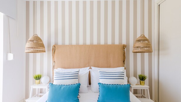 A bedroom with a striped wall, a large bed with two pillows, two bedside tables, and two hanging lamps.