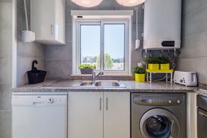 A modern kitchen with a built-in dishwasher, washing machine, and a window with a view of buildings.