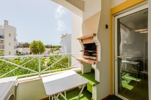 A balcony with a grill, a table, and a view of the neighborhood.