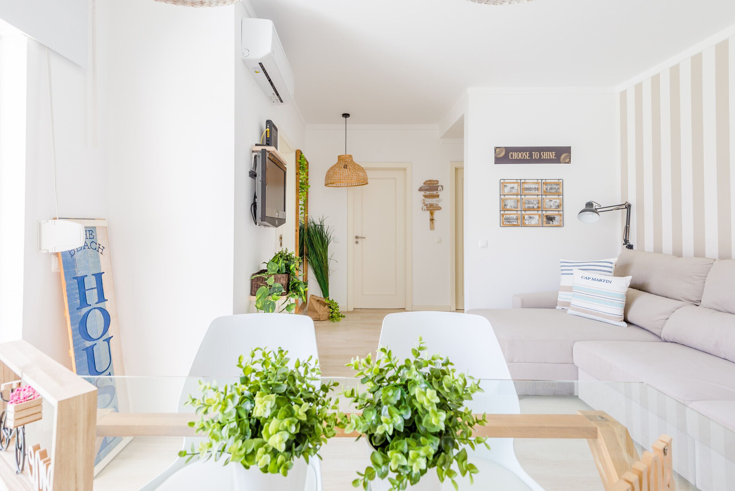 A modern living room with a sofa, a glass coffee table with plants, a TV, and a sign that says 'CHOOSE TO SHINE'.