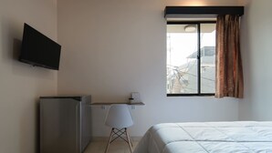 Une chambre à coucher avec un lit, un bureau avec une chaise, un téléviseur fixé au mur et une fenêtre avec des rideaux.