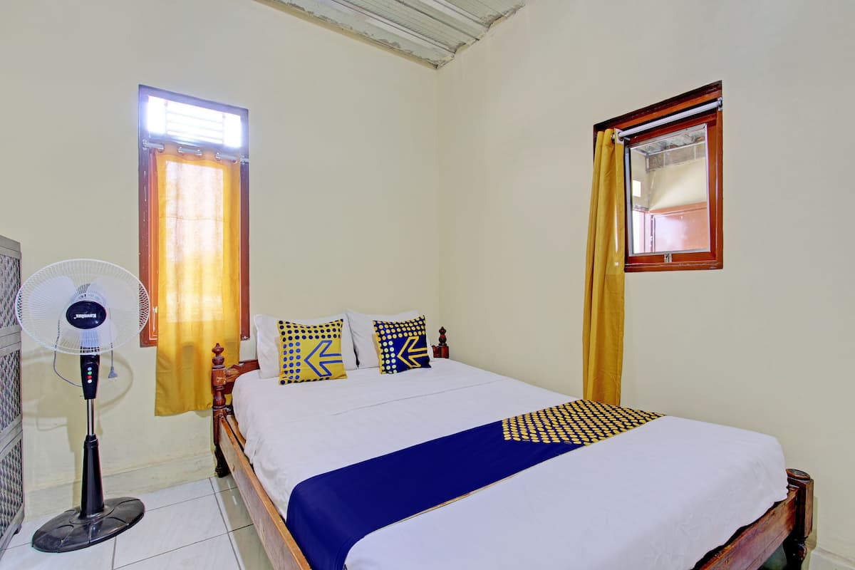 A bedroom with a bed, a standing fan, and two windows with curtains.