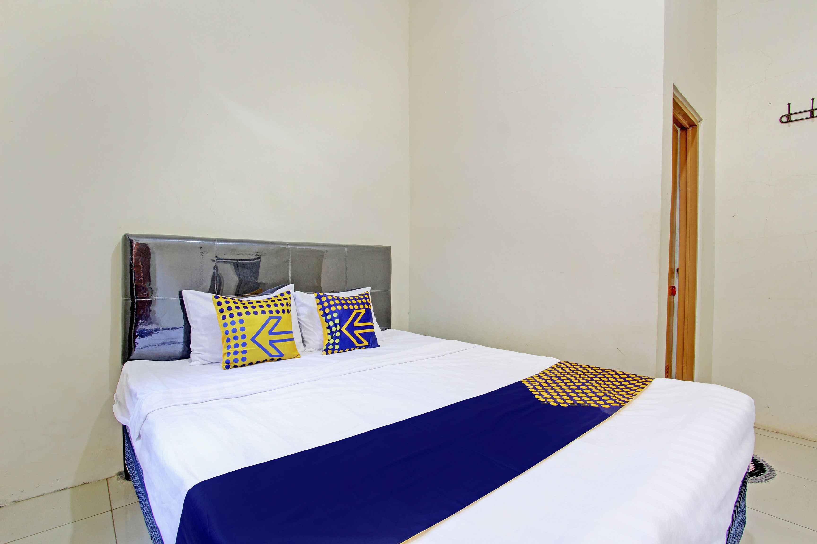 Una cama bien tendida con una colcha blanca y azul, dos almohadas con diseños geométricos, y una puerta de madera al fondo.