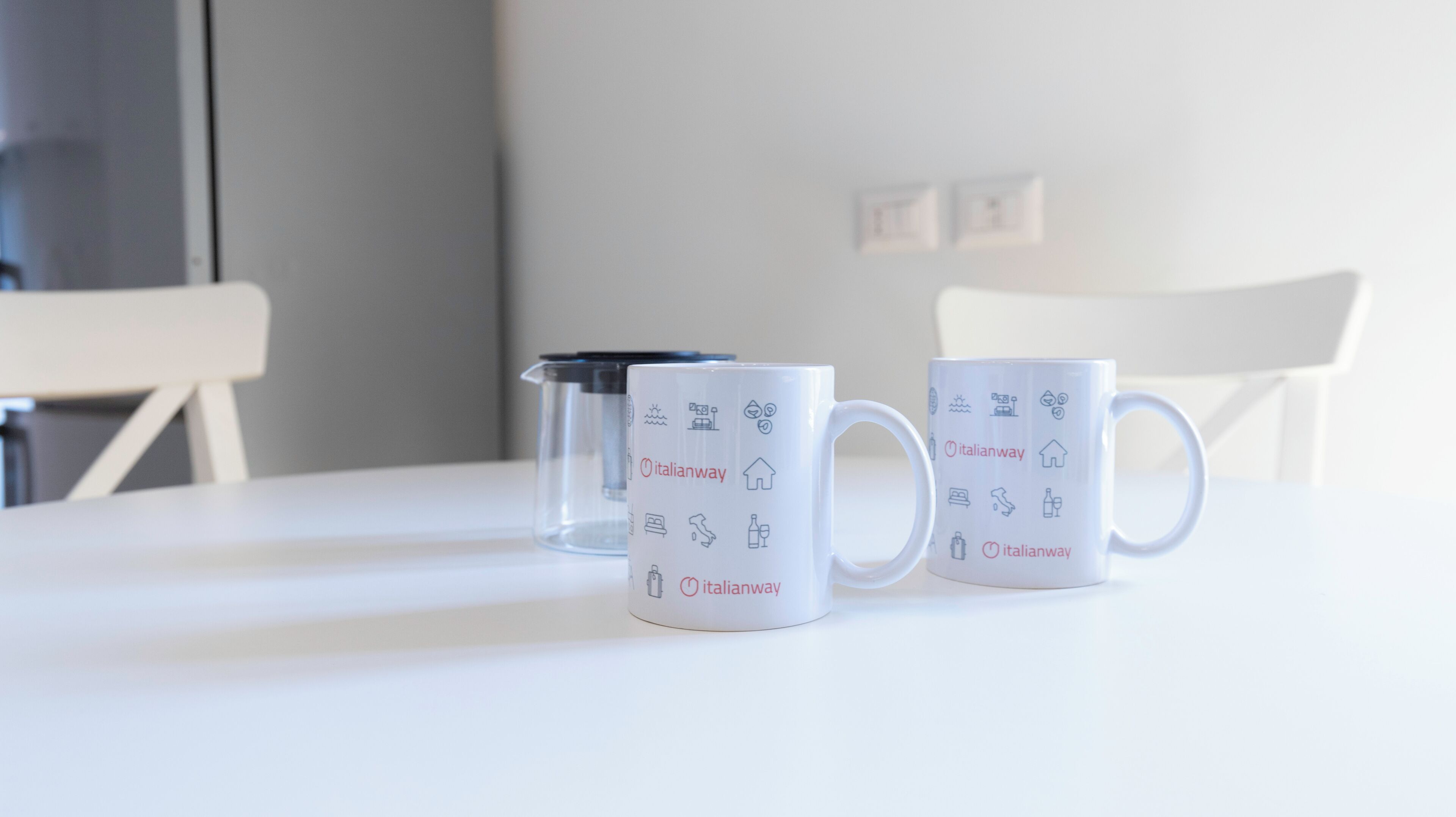 Two white mugs with Italianway logos and icons, placed on a white table.